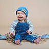 Malo a rejoint le concours — aidez-le/la à gagner de superbes lots ! baby, denim, overalls, blue_hat, smiling, sitting, beige_background, fabric, child, cute, happy, portrait, infant, clothing, studio, casual, feet, striped_shirt, person, toddler