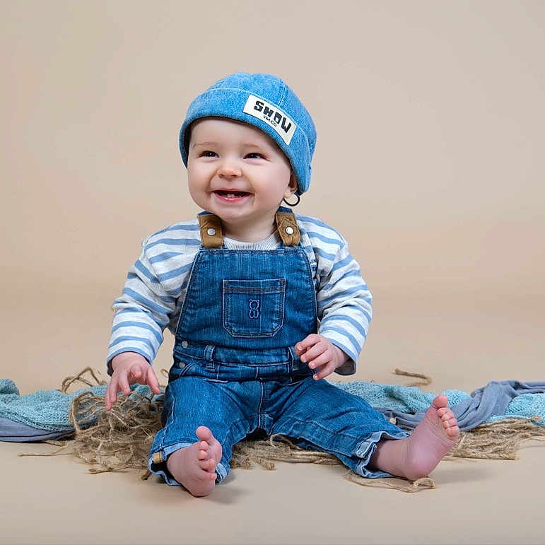 Malo a rejoint le concours — aidez-le/la à gagner de superbes lots ! baby, denim, overalls, blue_hat, smiling, sitting, beige_background, fabric, child, cute, happy, portrait, infant, clothing, studio, casual, feet, striped_shirt, person, toddler