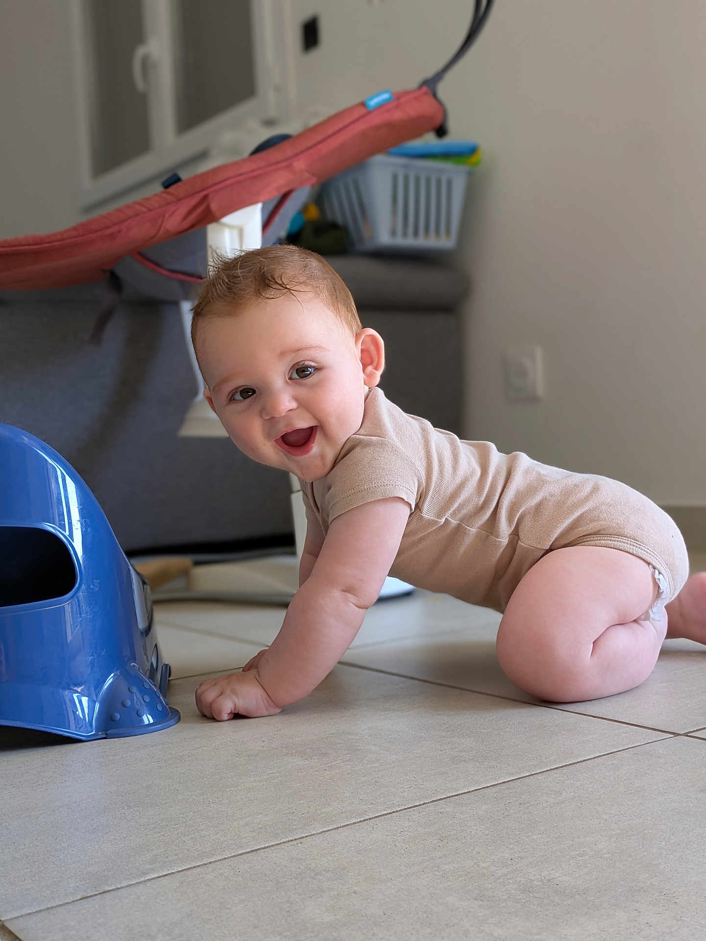 Nathan participe au concours pour gagner de l'argent avec cette photo : baby, crawling, onesie, indoor, floor, smiling, happy, child, tile_floor, blue_object, baby_bouncer, sofa, laundry_basket, home, cute, infant, person, young_child, playful, light