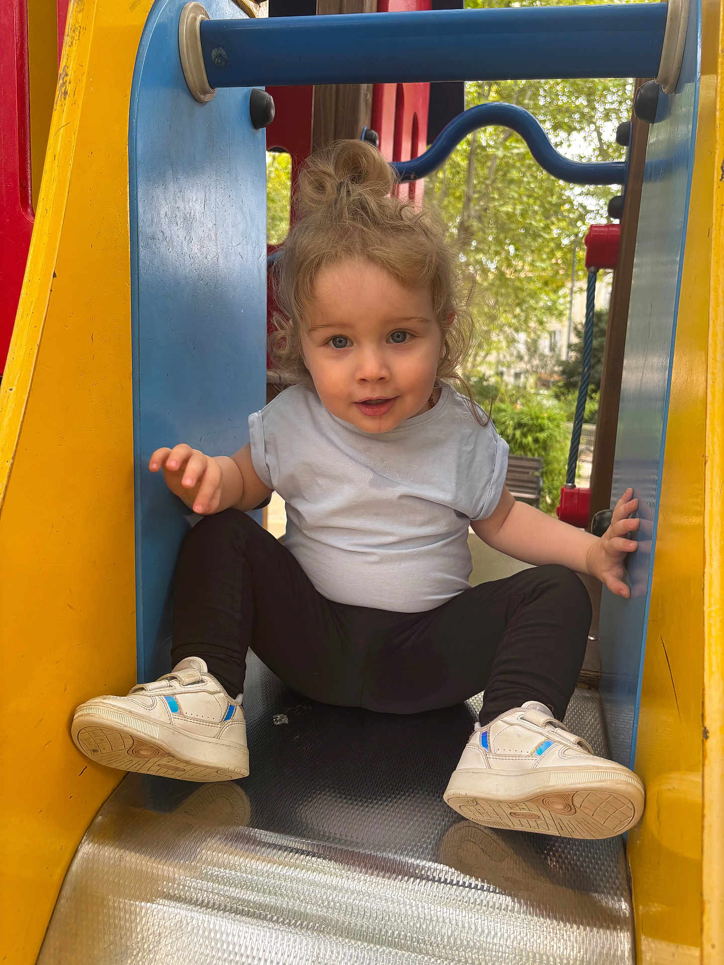 Shelly participe au concours pour gagner de l'argent avec cette photo : child, toddler, slide, playground, outdoor, sneakers, white_shirt, black_leggings, hair_bun, person, park, play_equipment, metal_slide, yellow, blue, red, nature, trees, daylight, smiling