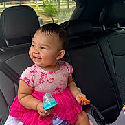 Khloe is registered to the contest to win money with this photo: baby, child, infant, smiling, sitting, car, car_interior, back_seat, pink_dress, dress, bottle, snack, toy, portrait, happy, face, indoors, daylight, automobile, seat