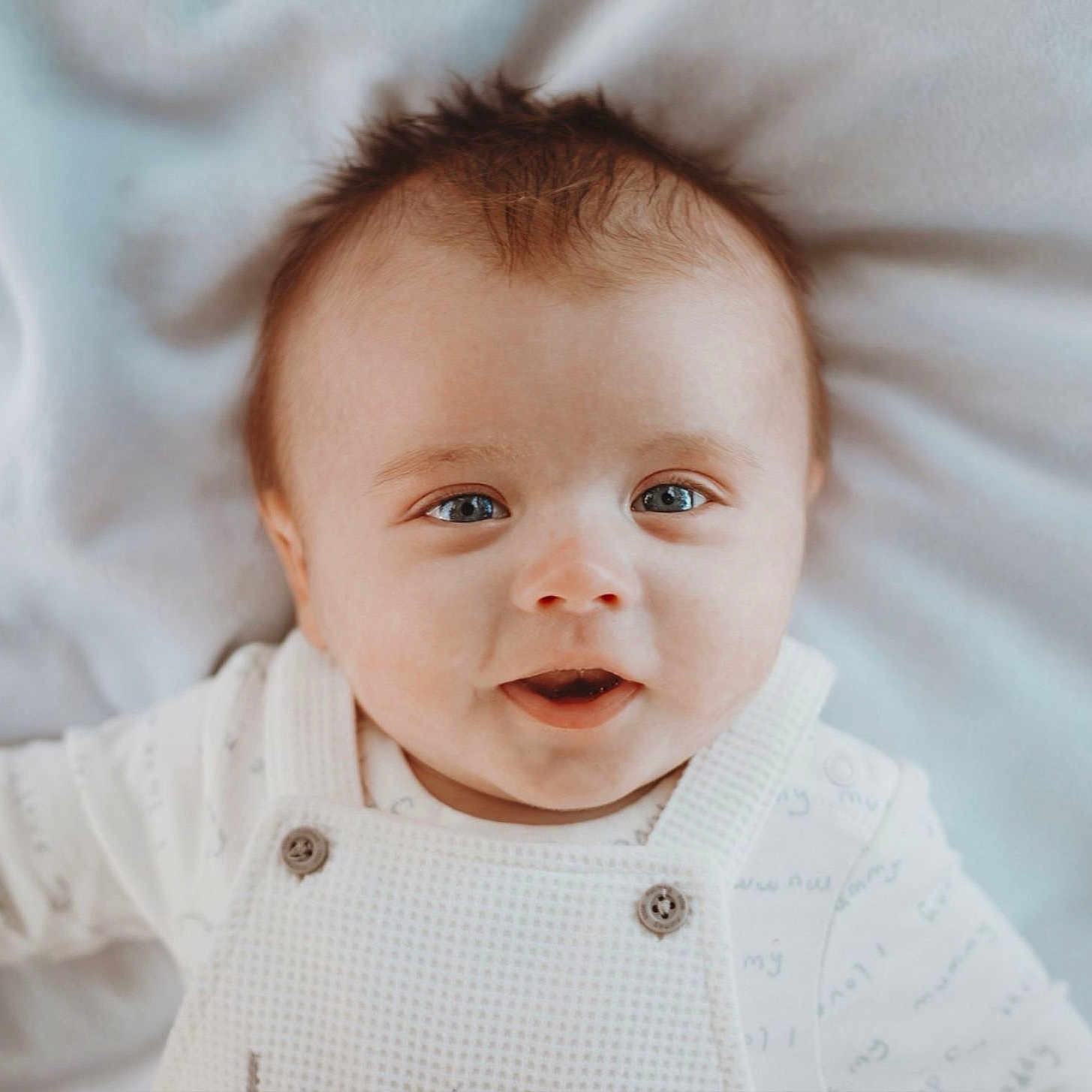 River-Bleu joined the competition — help win amazing prizes! baby, blanket, blue_eyes, buttoned_overalls, child, closeup, cute, expression, face, happy, head, indoors, infant, laying_down, portrait, skin, smiling, soft_lighting, white_clothing, young