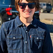 Bryler is registered to the contest to win money with this photo: child, young_person, denim_jacket, sunglasses, smile, outdoor, sunlight, casual, portrait, daytime, vehicle, tree, street, fashion, person, happy, cool, short_hair, blue, background_blur