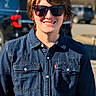 child, young_person, denim_jacket, sunglasses, smile, outdoor, sunlight, casual, portrait, daytime, vehicle, tree, street, fashion, person, happy, cool, short_hair, blue, background_blur