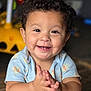 child, toddler, smile, clapping, curly_hair, blue_clothing, onesie, hot_air_balloon_pattern, happy, indoors, wooden_floor, face, baby, cute, hands, person, infant, portrait, sitting, playful