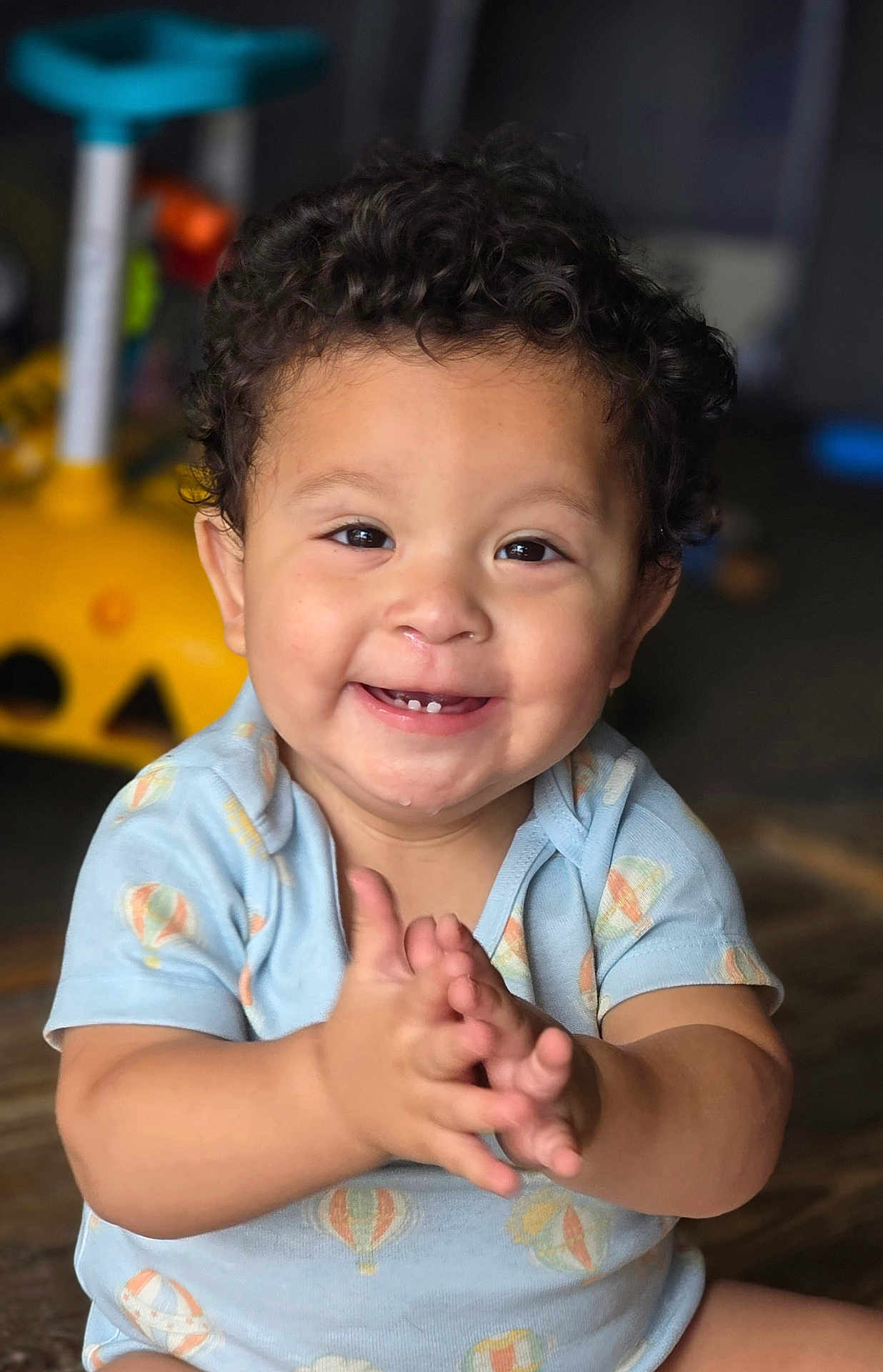 Lincoln is registered to the contest to win money with this photo: child, toddler, smile, clapping, curly_hair, blue_clothing, onesie, hot_air_balloon_pattern, happy, indoors, wooden_floor, face, baby, cute, hands, person, infant, portrait, sitting, playful