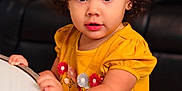 Ainhoa joined the competition — help win amazing prizes! toddler, child, yellow_dress, curly_hair, yellow_bow, table, indoor, living_room, portrait, cute, holding, big_eyes, flower_pattern, jewelry, furniture, smile, person, young_child, standing, curiosity