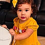 toddler, child, yellow_dress, curly_hair, yellow_bow, table, indoor, living_room, portrait, cute, holding, big_eyes, flower_pattern, jewelry, furniture, smile, person, young_child, standing, curiosity