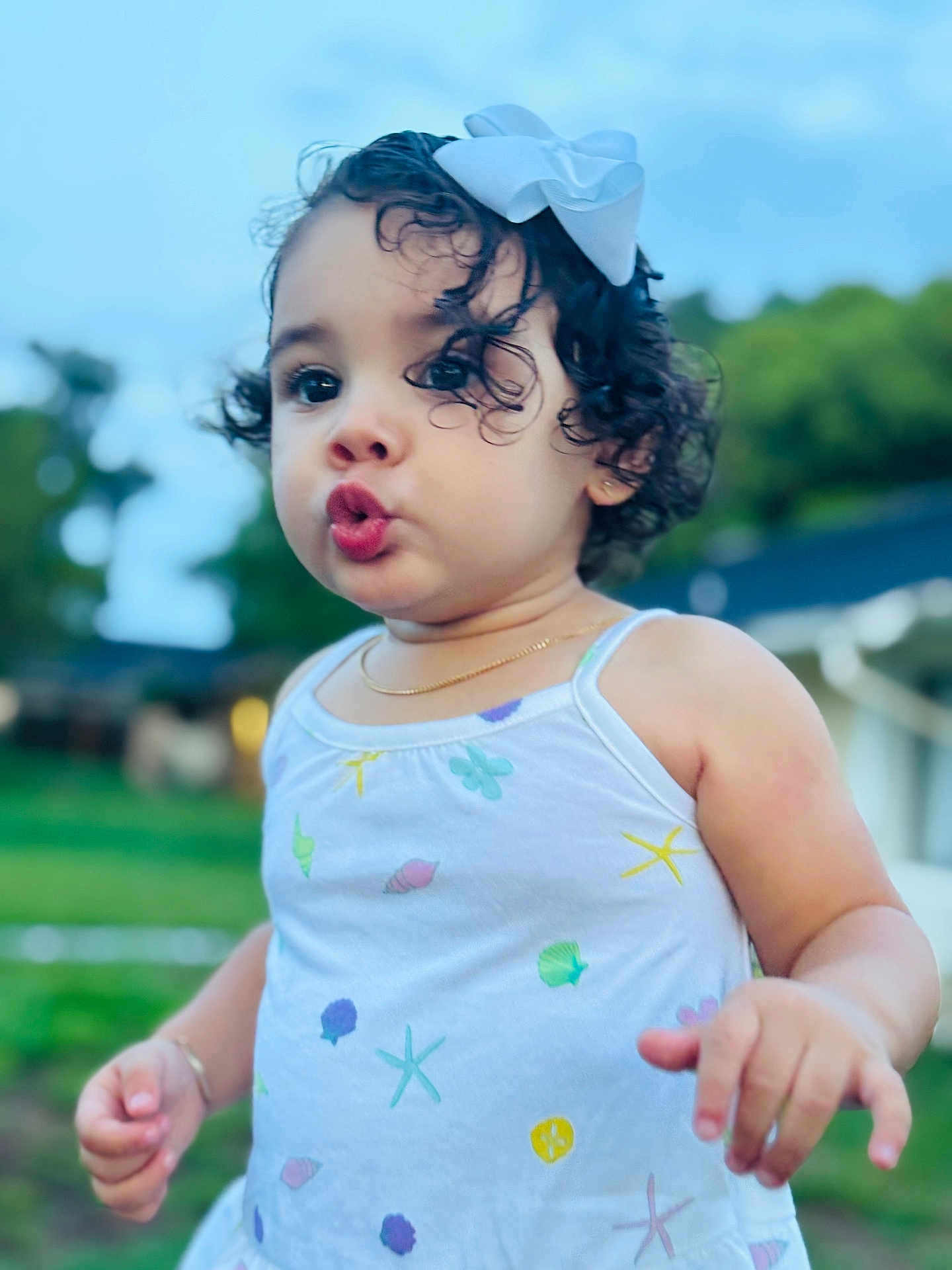 Ainhoa is registered to the contest to win money with this photo: child, toddler, curly_hair, bow, dress, outdoor, funny_face, expression, sea_shell_pattern, green_background, blue_sky, person, portrait, cute, summer, nature, jewelry, bracelet, gold_necklace, daylight