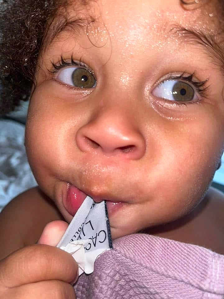 Neko Pitts joined the competition — help win amazing prizes! child, close_up, face, curly_hair, eyes, expression, hand, fabric, label, chewing, skin, cute, portrait, indoors, baby, toddler, curiosity, soft_texture, playful, young