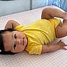 baby, infant, yellow_onesie, flower_hair_clip, pink_mattress, playpen, cute, lying_down, chubby_arms, chubby_legs, curious_eyes, indoor, soft_lighting, child, adorable, smiling, portrait, baby_clothing, head, hand