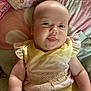 baby, infant, child, smiling, yellow_dress, lace, bed, blanket, floral_pattern, polka_dots, cute, chubby_cheeks, skin, head, arm, lying_down, indoors, soft_lighting, playful, tongue_out