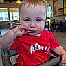 baby, child, infant, portrait, restaurant, high_chair, red_shirt, sitting, face, blue_eyes, brown_hair, smiling, hand, fingers, food, plastic_bag, indoor, furniture, background, photography