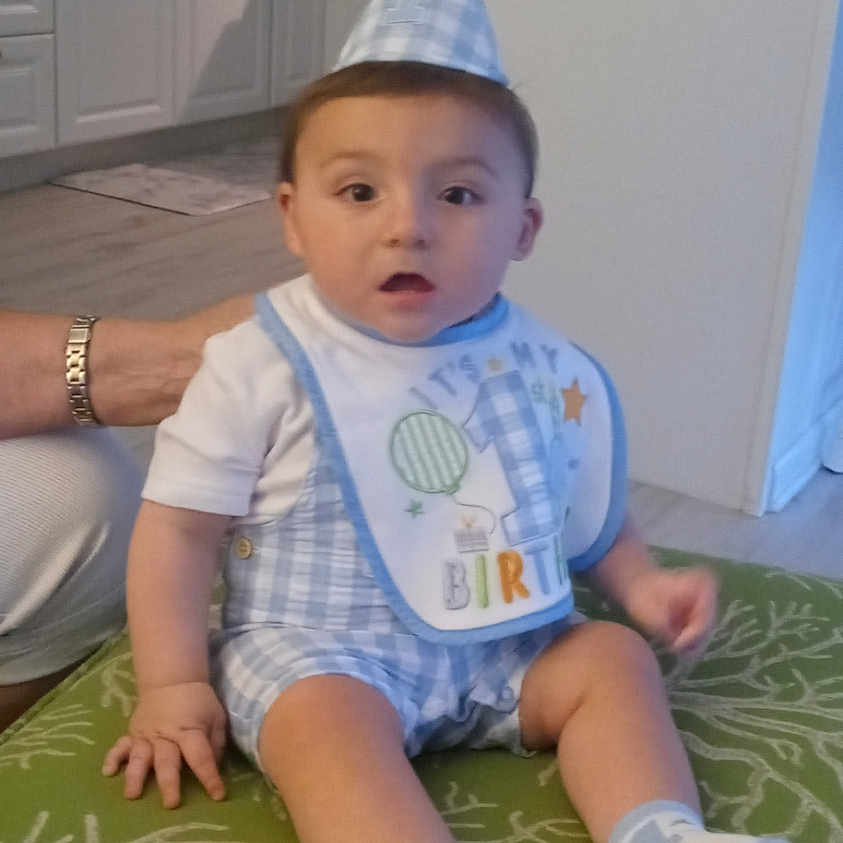 Liam is registered to the contest to win money with this photo: baby, birthday_hat, bib, blue_clothing, child, cute, floor, green_rug, indoor, kitchen, person, sitting, sock, white_cabinet, young_child, adult_arm, patterned_rug, curious_expression, casual, home_interior