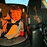 child, car_seat, sunlight, blue_eyes, car_interior, seatbelt, foot, red_ball, baby, portrait, seat, car_window, safety_harness, infant, cute, young_child, car_trip, sunset, casual_clothing, curious