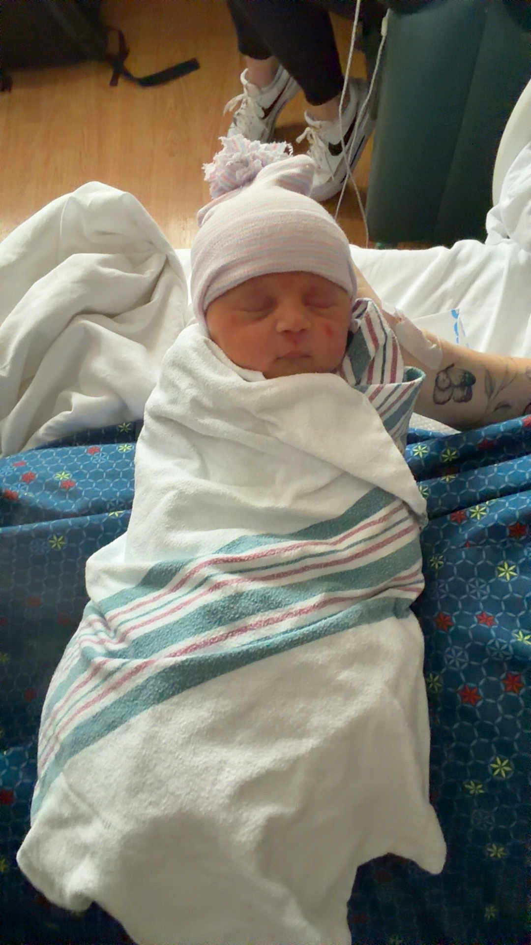 Isla is registered to the contest to win money with this photo: baby, newborn, swaddle, hospital_room, beanie, pom_pom_hat, sleeping, blanket, sheet, tattoo, arm, sneakers, shoes, hardwood_floor, bed, cozy, candid, portrait, striped_blanket, person