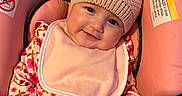 Celeste joined the competition — help win amazing prizes! baby, child, smiling, pink, hat, bib, car_seat, floral_pattern, knitted, pom_pom, cute, infant, clothing, portrait, happy, seated, indoors, soft_lighting, closeup, person