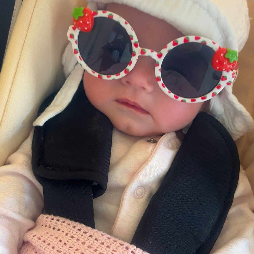 Eloise is registered to the contest to win money with this photo: accessory, baby, blanket, car_seat, child, clothing, cute, face, fashion, hat, headwear, indoors, infant, person, pink, portrait, seatbelt, sunglasses, warm, white