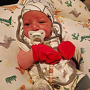 Ryatt joined the competition — help win amazing prizes! baby, newborn, infant, pacifier, pacifier_clip, mittens, red_mitten, hat, beanie, pillow, dinosaur_print, blanket, onesie, clothing, bead_chain, clip, closeup, portrait, indoors, baby_bedding