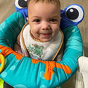 Zayden joined the competition — help win amazing prizes! baby, child, smiling, face, bib, baby_seat, colorful, blue, orange, green, indoor, floor, toy, cute, portrait, young_child, happy, clothing, person, infant