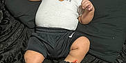 Binyamyin is registered to the contest to win money with this photo: baby, infant, child, person, face, clothing, onesie, shorts, sneakers, nike, basketball_logo, black_blanket, patterned_blanket, lying_down, indoor, cute, chubby_cheeks, wide_eyes, footwear, sportswear
