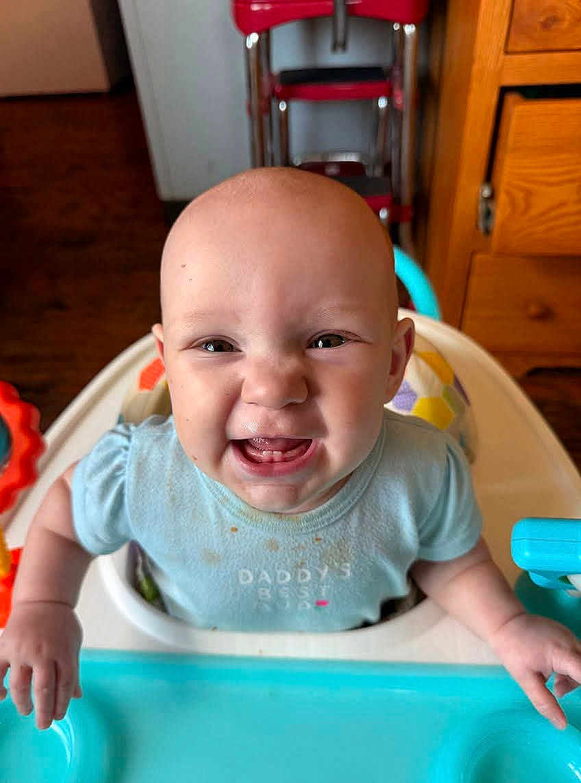 Ruth joined the competition — help win amazing prizes! baby, high_chair, smiling, expression, indoor, child, furniture, wood_floor, toy, blue_shirt, face, hands, mouth_open, teeth, kitchen, cabinet, colorful, person, cute, happy