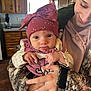 Ruth is registered to the contest to win money with this photo: baby, child, woman, smile, hat, bow, knitted_clothing, hand, holding, indoor, kitchen, cabinet, hoodie, camo_jacket, person, face, expression, warm_lighting, home, cute