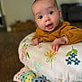 baby, infant, face, expression, cushion, floral_pattern, yellow_clothing, hand, crocheted_blanket, wooden_floor, indoors, person, child, portrait, curious, wide_eyes, cute, soft_light, home, blurred_background