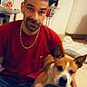 blur, bowl, brown_and_white_dog, casual, chain_necklace, dog, face, floor, furniture, home, indoor, man, pet, plant, red_shirt, short_hair, sitting, smile, table, tile_floor