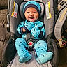 baby, infant, child, smiling, happy, car_seat, safety_harness, blue_outfit, hat, mittens, footed_pajamas, dinosaur_pattern, portrait, indoor, vehicle_interior, travel, seated, carpet, backpack, warning_label