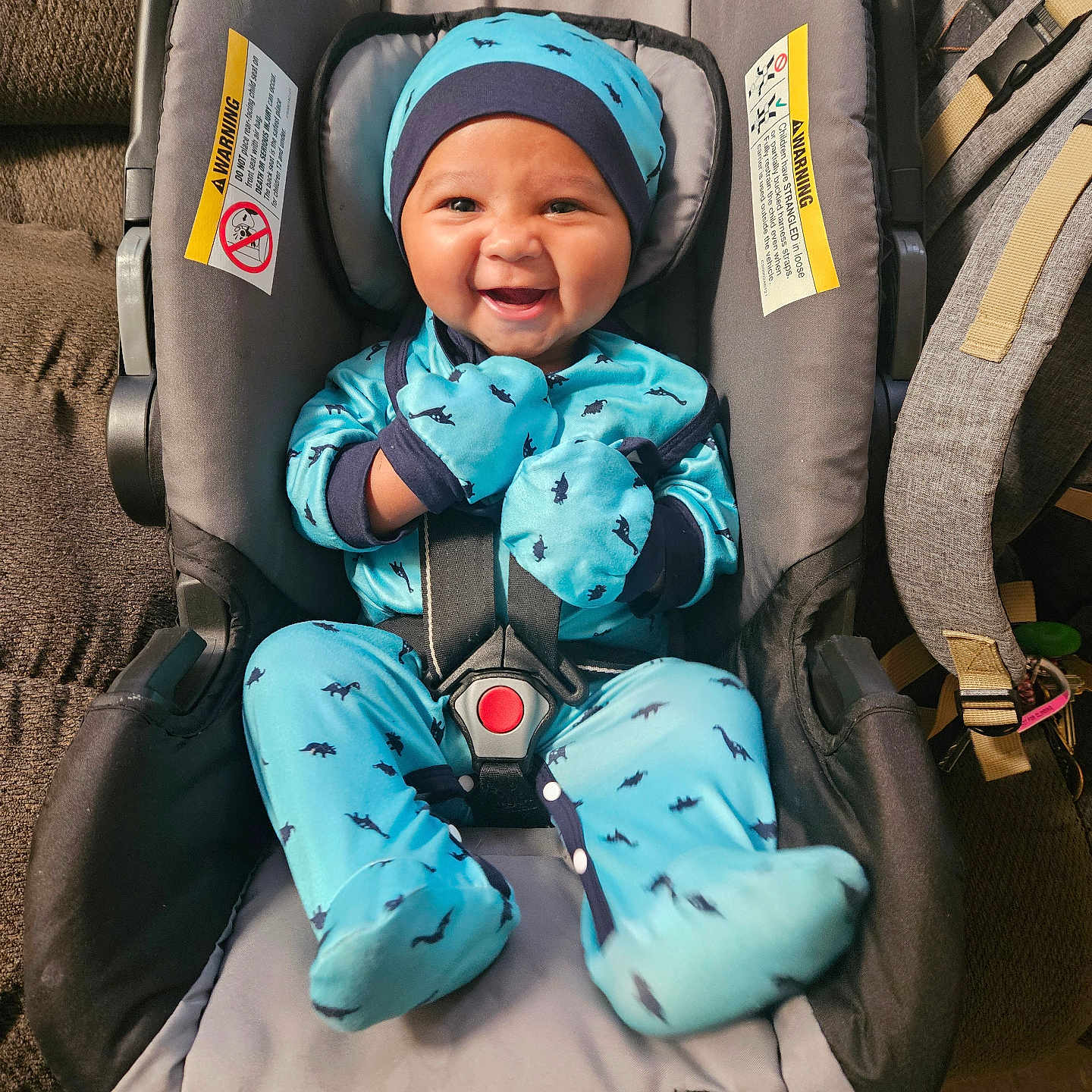 Joshua is registered to the contest to win money with this photo: baby, infant, child, smiling, happy, car_seat, safety_harness, blue_outfit, hat, mittens, footed_pajamas, dinosaur_pattern, portrait, indoor, vehicle_interior, travel, seated, carpet, backpack, warning_label
