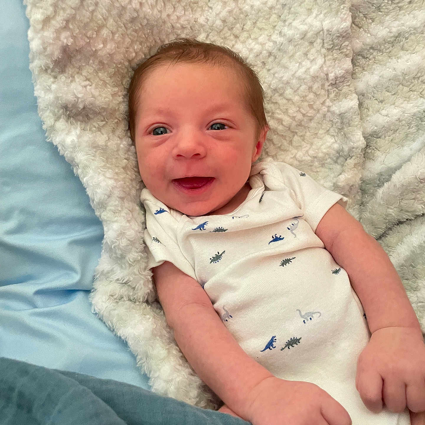 Johnathon joined the competition — help win amazing prizes! baby, bed, blanket, child, cozy, cute, dinosaur_pattern, face, hands, happy, indoor, infant, lying_down, newborn, onesie, person, portrait, skin, smiling, soft