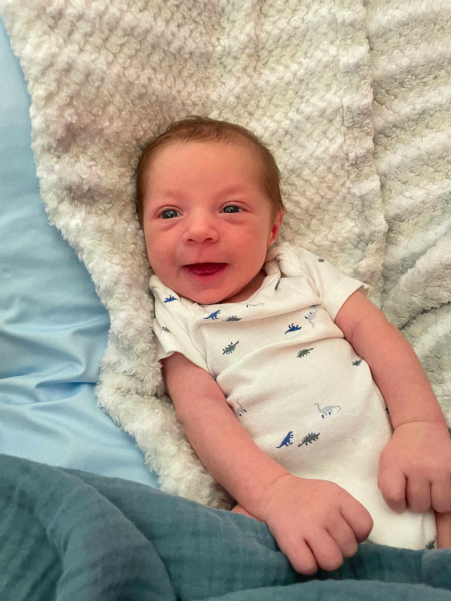 Johnathon joined the competition — help win amazing prizes! baby, newborn, smiling, blanket, onesie, dinosaur_pattern, infant, cozy, soft, cute, hands, face, skin, lying_down, bed, happy, child, person, indoor, portrait