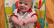 Noah is registered to the contest to win money with this photo: baby, infant, smile, baby_seat, colorful, patterned, wooden_floor, clothing, happy, child, indoor, seat_belt, cute, portrait, person, young_child, sitting, hands, face, caucasian