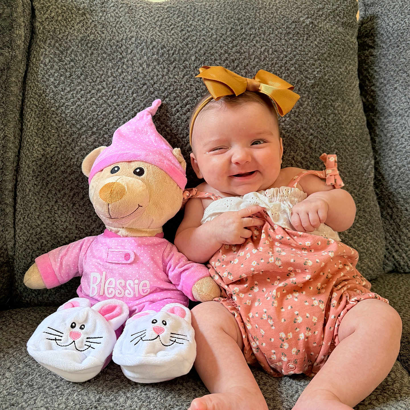 Blessie Rosalie is registered to the contest to win money with this photo: baby, bow, smile, plush_toy, teddy_bear, pink_clothing, romper, couch, gray_couch, indoor, child, cute, happy, sitting, footwear, bunny_slippers, headband, toy, infant, cozy
