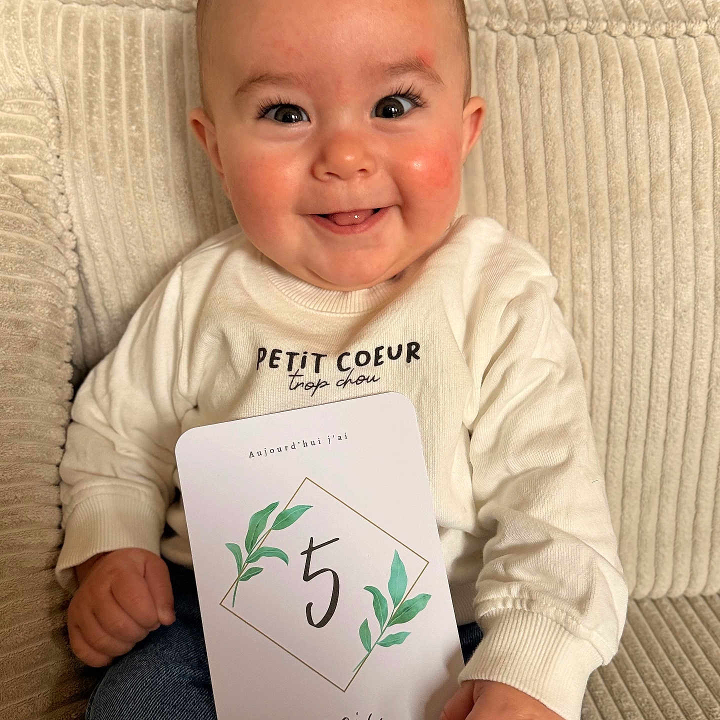 Noah participe au concours pour gagner de l'argent avec cette photo : baby, smile, infant, milestone_card, couch, sweater, jeans, happy, face, hand, indoor, child, person, portrait, seated, cute, skin, eyelashes, background, texture