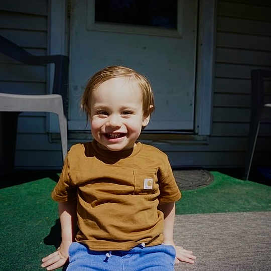 Colt joined the competition — help win amazing prizes! child, toddler, smiling, outdoor, door, plastic_chair, green_carpet, sunlight, casual_clothing, happy, sitting, brown_shirt, blue_pants, front_yard, shadow, person, daylight, home, porch, young_child