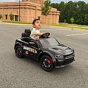 Daniell is registered to the contest to win money with this photo: toddler, child, toy_car, black_car, parking_lot, outdoor, person, vehicle, cute, boy, small_car, asphalt, tree, building, greenery, daytime, looking_up, play, fun, casual
