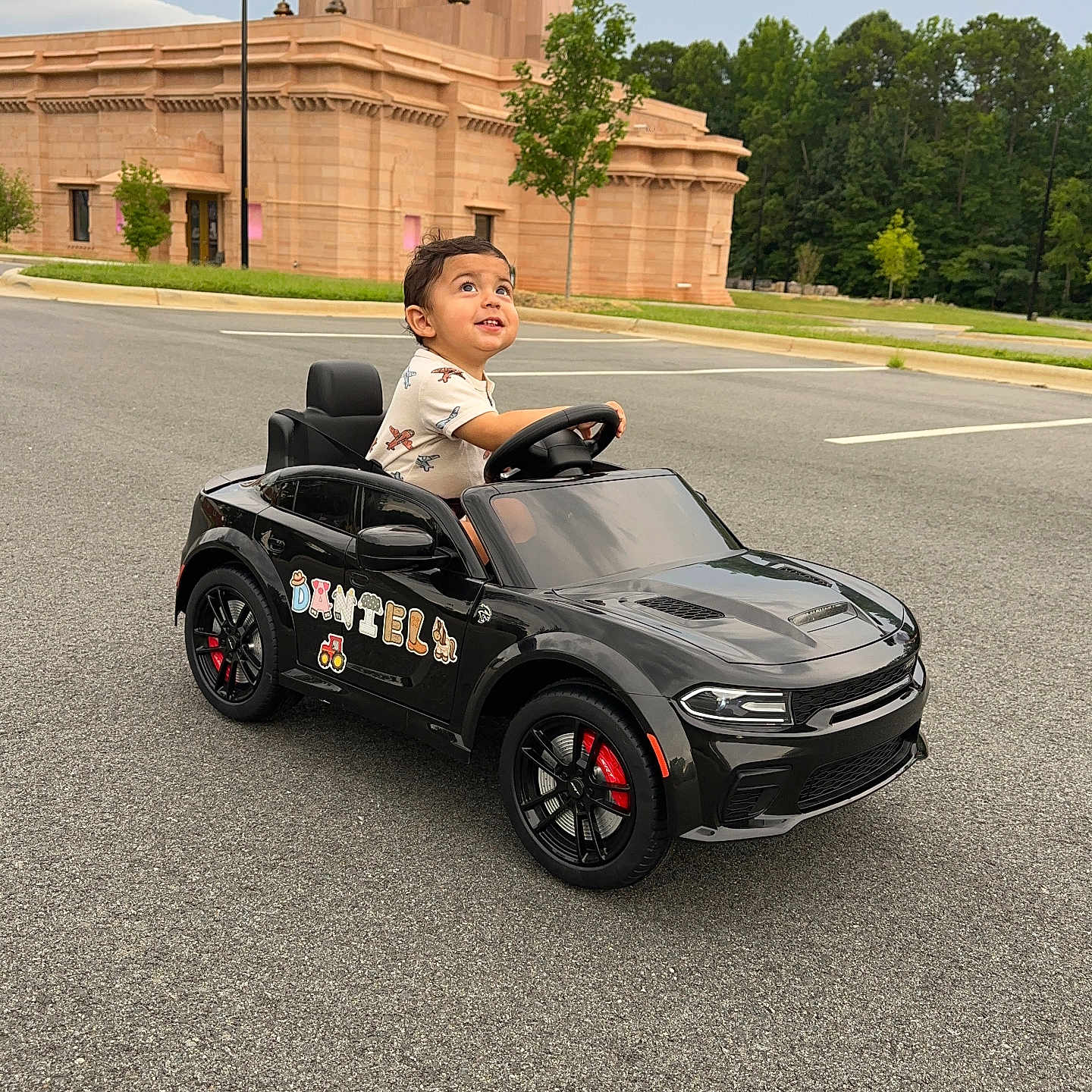 Daniell is registered to the contest to win money with this photo: asphalt, black_car, boy, building, casual, child, cute, daytime, fun, greenery, looking_up, outdoor, parking_lot, person, play, small_car, toddler, toy_car, tree, vehicle