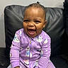 baby, child, smiling, laughing, purple_onesie, animal_prints, toy, red_toy, sitting, black_leather_couch, indoor, happy, cute, infant, young_child, face, expression, clothing, joyful, portrait
