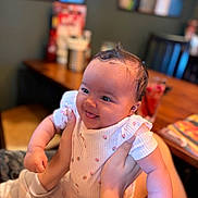 Alaiyah joined the competition — help win amazing prizes! baby, infant, smiling, happy, tongue_out, hands, indoor, table, chair, clothing, patterned_outfit, person, face, cute, joyful, skin, hair, holding, blurred_background, warm_light
