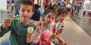 Ines a rejoint le concours — aidez-le/la à gagner de superbes lots ! child, children, ice_cream, cone, bench, shopping_mall, indoor, people, casual_clothing, curly_hair, smiling, playful, food, dessert, retail, storefront, public_space, family, leisure, happy