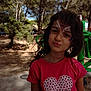child, girl, face_paint, red_shirt, heart_design, outdoor, trees, nature, playful, portrait, smile, daylight, hair, expression, park, young, casual_clothing, sunlight, green_equipment, background