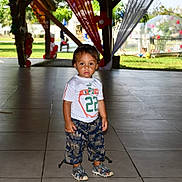 Manoa participe au concours pour gagner de l'argent avec cette photo : child, toddler, standing, pavilion, balloons, decorations, outdoor, clothing, sandals, tile_floor, curtains, grass, trees, daylight, person, number_22, white_shirt, patterned_pants, serious_face, event