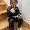 Manoa a rejoint le concours — aidez-le/la à gagner de superbes lots ! toddler, child, stairs, pacifier, black_jacket, white_shoes, indoors, wooden_stairs, casual_clothing, sitting, young_child, curious_expression, white_wall, footwear, holding_object, portrait, person, hair, face, comfortable