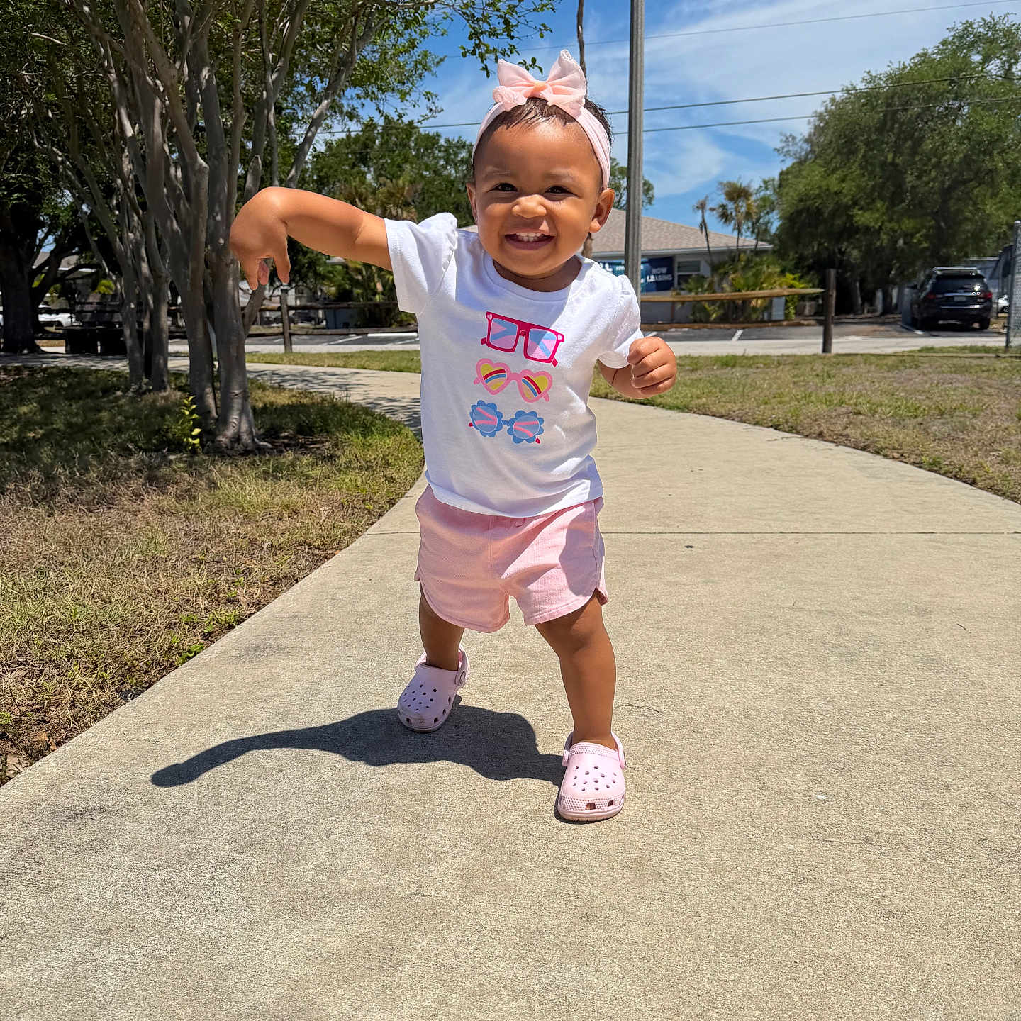 Teyani joined the competition — help win amazing prizes! toddler, child, outdoor, sidewalk, sunny, blue_sky, trees, grass, pink_bow, white_tshirt, pink_shorts, pink_shoes, happy, smiling, walking, person, clothing, daylight, nature, portrait