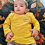 baby, child, yellow_shirt, lying_down, blanket, floral_pattern, curious_expression, indoors, portrait, young_child, clothing, hand, face, head, person, soft_lighting, casual_wear, cute, small_hands, comfortable