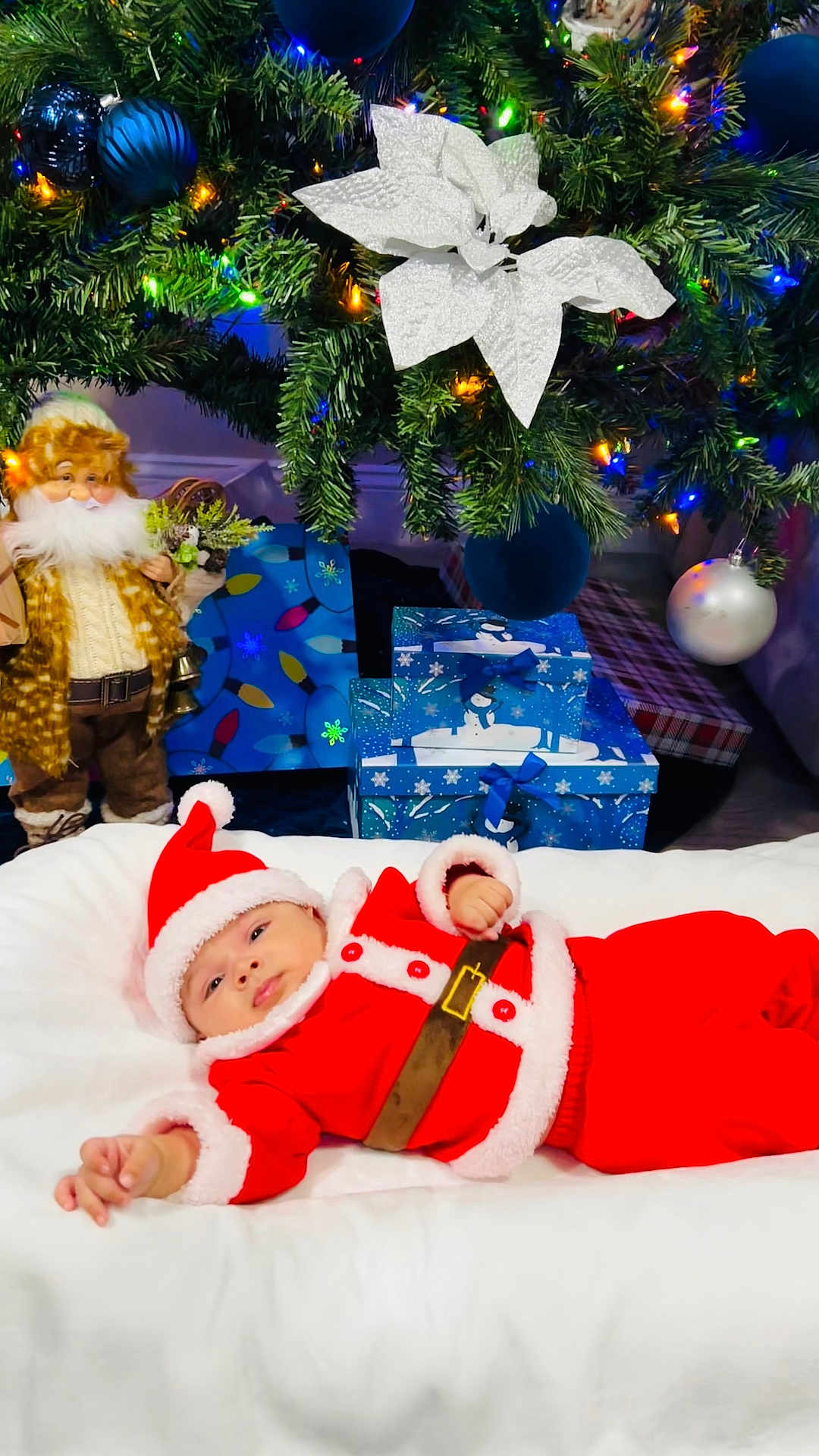 Nathan is registered to the contest to win money with this photo: baby, christmas_tree, christmas_lights, ornaments, gift_boxes, santa_claus_outfit, holiday, festive, doll, white_blanket, red_clothing, beard, decorations, blue_ornaments, indoor, celebration, toddler, holiday_spirit, winter, festivity
