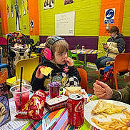 Briar is registered to the contest to win money with this photo: child, ear_protection, sandwich, snacks, coca_cola, fries, table, colorful, indoor, play_area, cartoon_wall_decals, person, phone, chair, stroller, juice, cup, food, casual, jacket