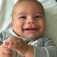 Matheus is registered to the contest to win money with this photo: baby, smiling, close_up, infant, hands, clapping, gray_clothing, cheeks, happy, face, portrait, young_child, cute, indoor, blanket, soft_light, expression, head, skin, person